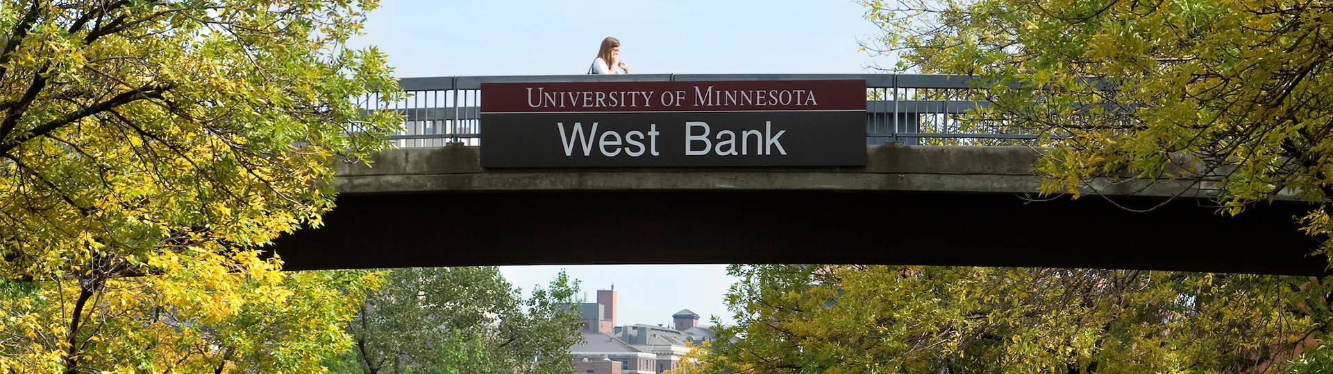 A student walks across a footpath with a sign that reads "University of Minnesota West Bank"
