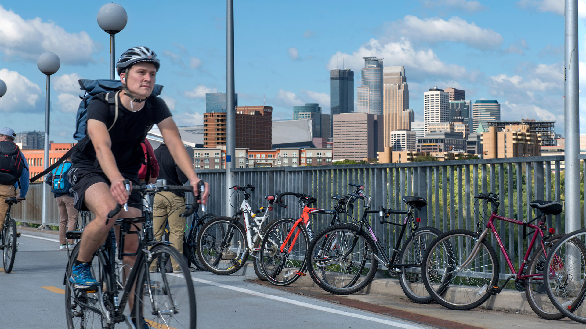 A student in a black shirt and shorts bikes down the Washington Avenue Bridge, with the Minneapolis skyline in the background